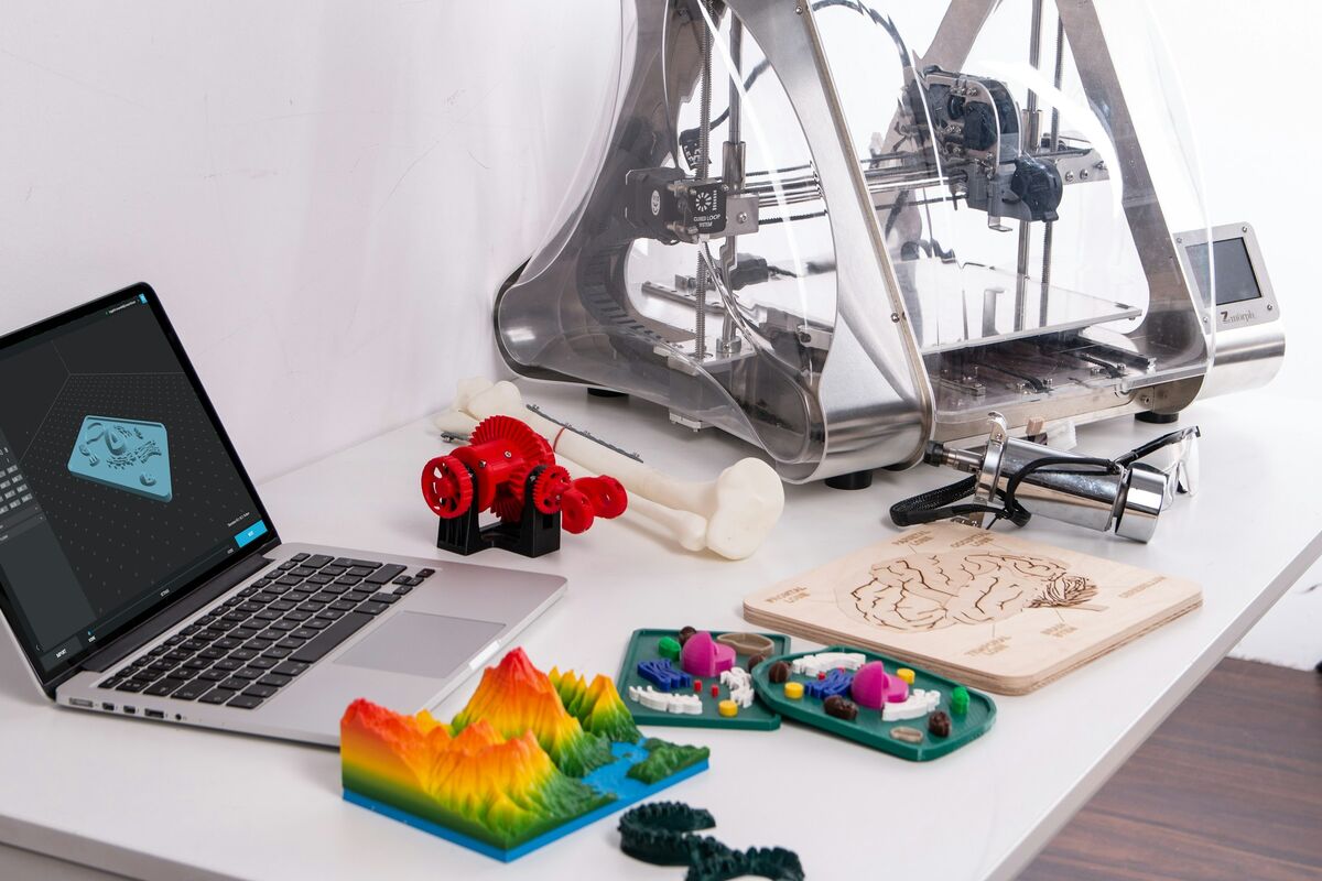 3D tiskárna, notebook s modelem a různé barevné výtisky na stole